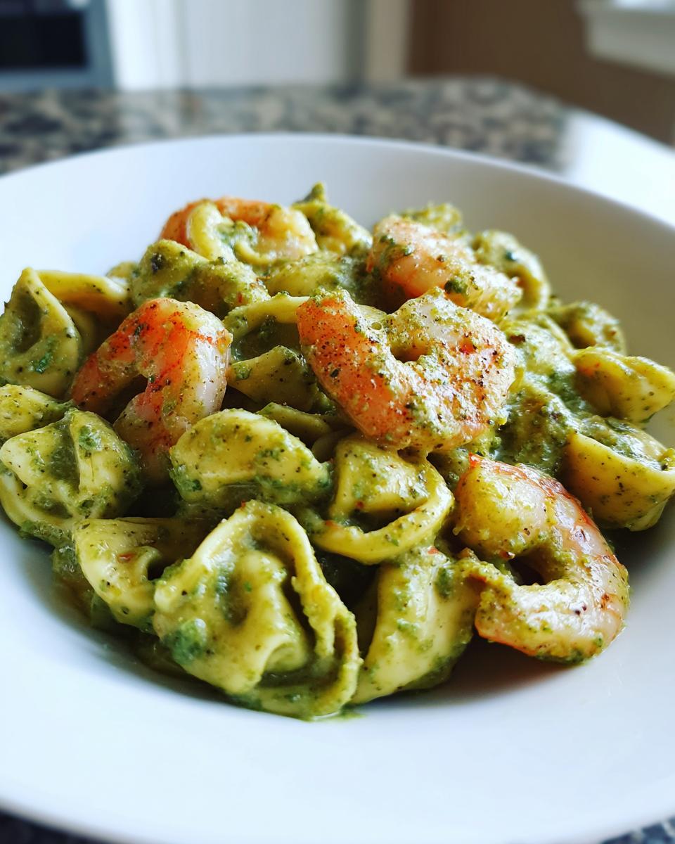 A close-up of a white bowl filled with Creamy Shrimp Pesto Tortellini Pasta, featuring plump shrimp and green pesto sauce.