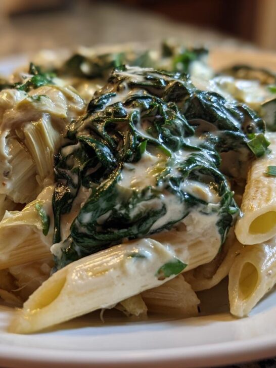 Close-up of a serving of Creamy Spinach Artichoke Penne Pasta topped with rich sauce, spinach, and artichoke hearts.