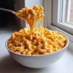 A spoonful of incredibly cheesy and creamy stovetop Southern mac cheese being lifted from a white bowl, showing long cheese pulls.