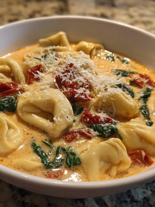 A close-up of a white bowl filled with Creamy Tuscan Tortellini Soup, featuring tortellini, spinach, sun-dried tomatoes, and grated Parmesan.