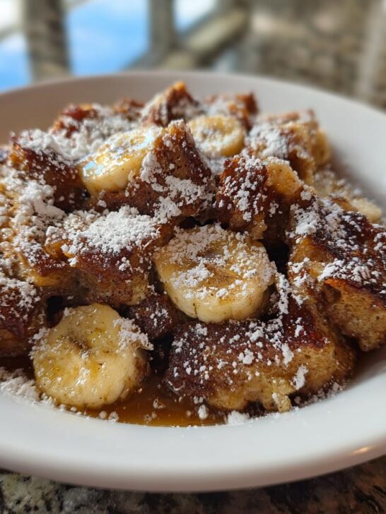 A white bowl filled with cubes of Crock Pot Creamy Banana French Toast, topped with banana slices and powdered sugar.