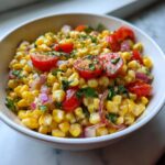 Close-up of Easy Loaded Corn Salad featuring bright yellow corn, red cherry tomato halves, and purple onion in a white bowl.