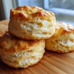 Three tall, golden-brown Flaky Honey Buttermilk Biscuits stacked and resting on a wooden cutting board.