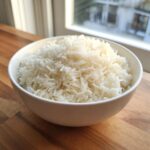 A white bowl filled with perfectly cooked, fluffy Basmati rice sitting on a wooden countertop near a bright window.