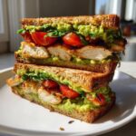 A tall, stacked Grilled Chicken Avocado Sandwich featuring grilled chicken, mashed avocado, and cherry tomatoes on toasted bread.