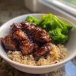 A bowl of Healthy Weeknight General Tsos Chicken coated in dark sauce, served over brown rice with steamed broccoli florets.