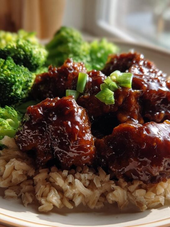 A plate featuring Healthy Weeknight General Tsos Chicken glazed in sauce, served over brown rice with steamed broccoli florets.