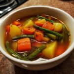 Close-up of a white bowl filled with rich broth Vegetable Soup featuring chunks of carrots, potatoes, and green beans.