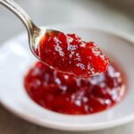 Close-up of a spoonful of glistening, bright red Homemade Strawberry Jam being lifted from a white bowl.