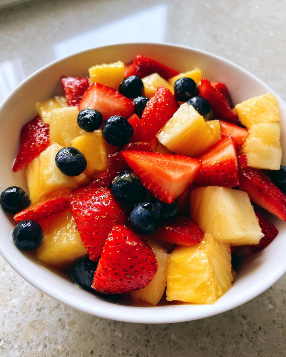 Close-up of a white bowl filled with Honey Lime Fruit Salad featuring strawberries, blueberries, and pineapple chunks.