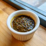 Close-up of thick, dark green Jamaican Jerk Seasoning Marinade with visible chili flakes in a white ramekin.
