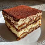 A close-up, square slice of layered Tiramisu showing coffee-soaked ladyfingers and creamy mascarpone filling, topped with cocoa powder.
