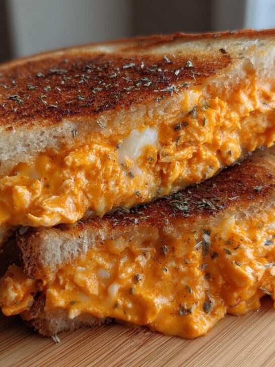 Two halves of My Favorite Buffalo Chicken Melt Sandwich stacked, showing gooey orange filling and toasted bread sprinkled with herbs.