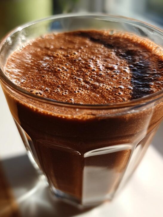 Close-up of a rich, dark brown Peanut Butter Cup Smoothie with frothy bubbles on top, sitting in a glass by a window.