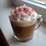 A clear glass mug filled with a Peppermint White Chocolate Mocha Latte, topped high with whipped cream and crushed candy canes.
