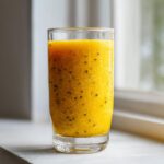 A vibrant yellow Pineapple Passion Fruit Smoothie in a tall glass, featuring visible black chia seeds.
