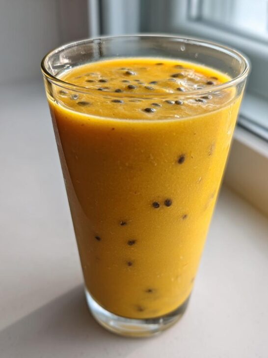 A tall glass filled with a thick, bright orange Pineapple Passion Fruit Smoothie topped with passion fruit seeds.