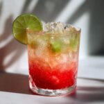 A cold, sweating glass filled with a layered Pineapple Watermelon Mocktail, ice, and a lime wheel garnish.
