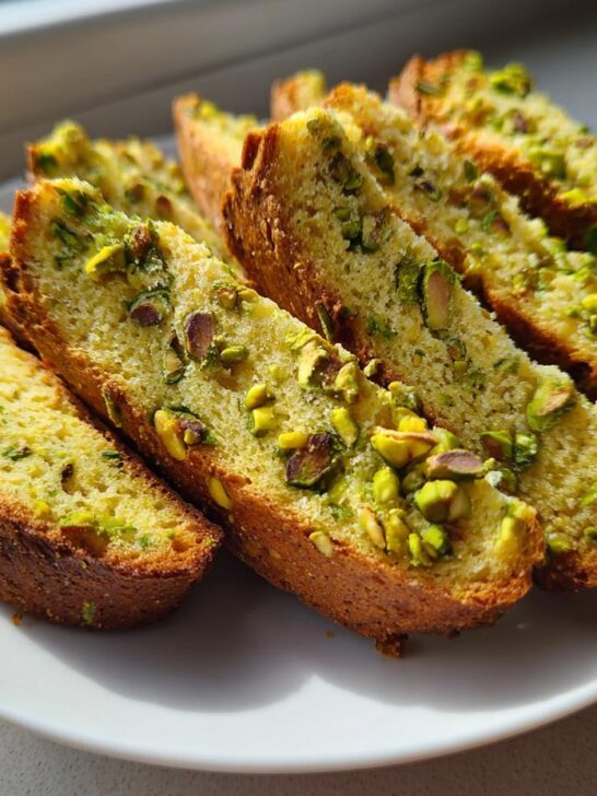 Several golden-brown slices of Pistachio Biscotti arranged on a white plate, featuring visible green pistachio pieces.