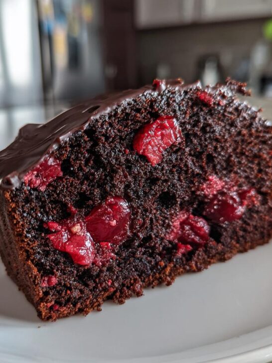 A moist slice of Quick Cherry Chocolate Cake featuring dark chocolate crumb and whole cherries, topped with chocolate ganache.