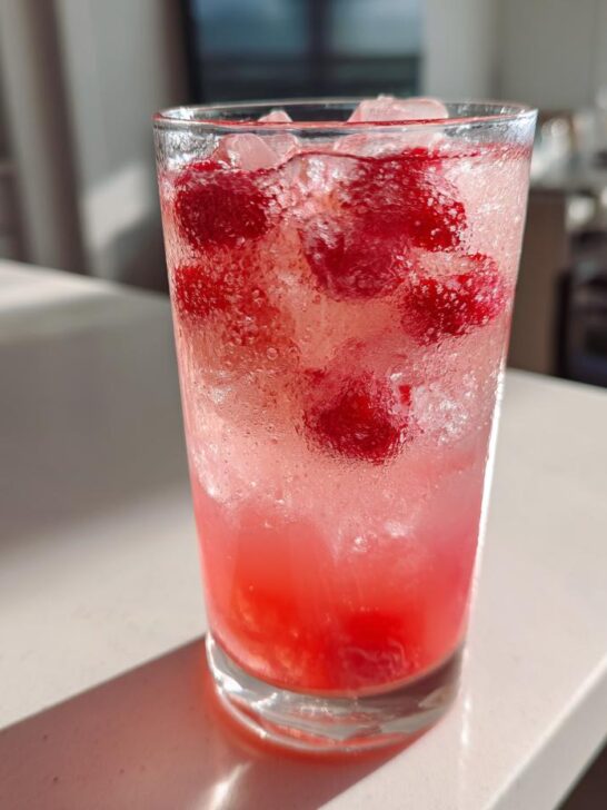 A tall, sweating glass filled with pink Raspberry Peach Lemonade, ice, and whole raspberries, sitting on a white counter.