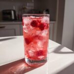 A tall glass filled with icy Raspberry Peach Lemonade, whole raspberries, and condensation on the glass.