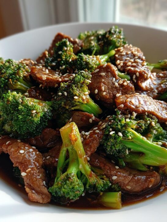A close-up of glistening Sesame Beef And Broccoli coated in a dark sauce and topped with white sesame seeds, served in a white bowl.