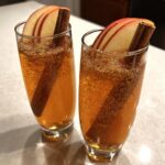 Two tall glasses filled with a sparkling Apple Cider Mocktail, garnished with apple slices and a cinnamon stick.