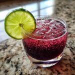 A close-up of a vibrant, dark red Yuletide Daisy Cocktail, garnished with a bright lime wheel, sitting on a granite counter.