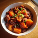 Close-up of a white bowl filled with rich, dark Spicy Vegan Jamaican Stew Peas featuring large chunks of orange pumpkin and topped with green onions.