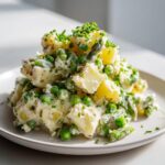 A mound of Spring Vegetable Potato Salad featuring chunks of potato, bright green peas, asparagus, and a creamy dressing, topped with fresh chives.