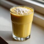 A vibrant yellow Tropical Pineapple Coconut Smoothie in a glass, topped with shredded coconut flakes.
