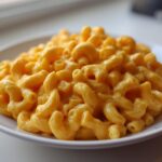 A close-up of a white plate piled high with creamy, bright orange macaroni pasta, representing The Ultimate Vegan Mac Cheese.