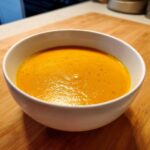 A close-up of a bowl of rich, orange Vegan Cheesy Carrot Soup, speckled with pepper, sitting on a wooden cutting board.