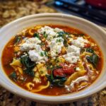 A close-up of a white bowl filled with rich tomato broth, pasta noodles, spinach, and topped with dollops of vegan ricotta for Vegan Lasagna Soup.