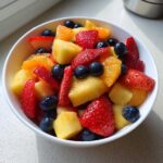 Close-up of a white bowl filled with colorful Honey Lime Fruit Salad featuring strawberries, pineapple, blueberries, and orange segments.