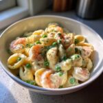 A close-up of White Cheddar Shells And Cheese With Shrimp And Peas served in a white bowl, garnished with herbs.