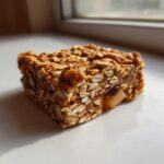 A single, chewy Apple Cinnamon Breakfast Bars square made with rolled oats, sitting on a white surface near a window.