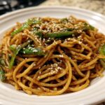 A close-up shot of a serving of glossy Asian Sesame Noodles topped with bright green scallions and white sesame seeds.