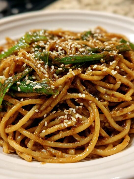 A close-up shot of a serving of glossy Asian Sesame Noodles topped with bright green scallions and white sesame seeds.