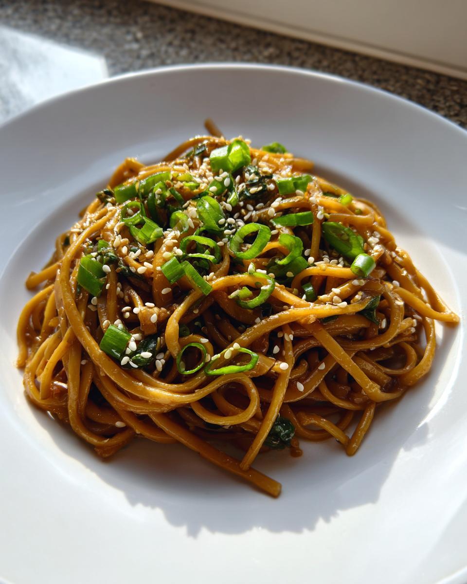 A close-up of a serving of glossy Asian Sesame Noodles topped with fresh green scallions and white sesame seeds on a white plate.