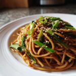 A close-up shot of glossy Asian Sesame Noodles topped with fresh green scallions and toasted sesame seeds.