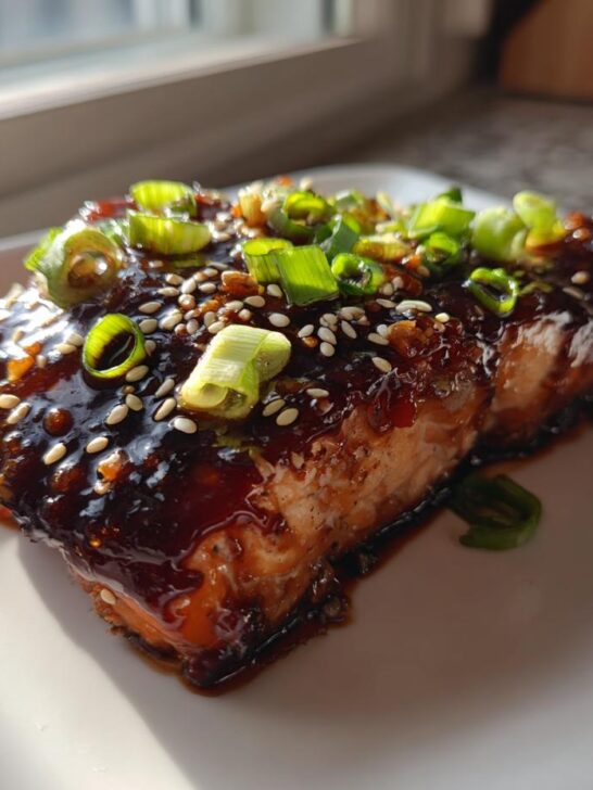 A close-up of a perfectly cooked piece of Baked Teriyaki Salmon covered in a thick, dark glaze, topped with sesame seeds and green onions.