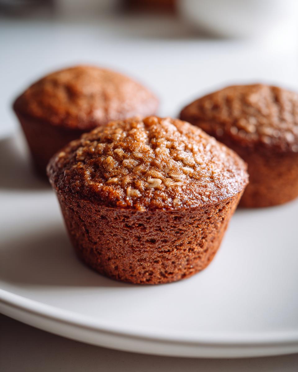 Three golden-brown Banana Oatmeal Muffins sitting on a white plate, with the front muffin in sharp focus showing the oat topping.