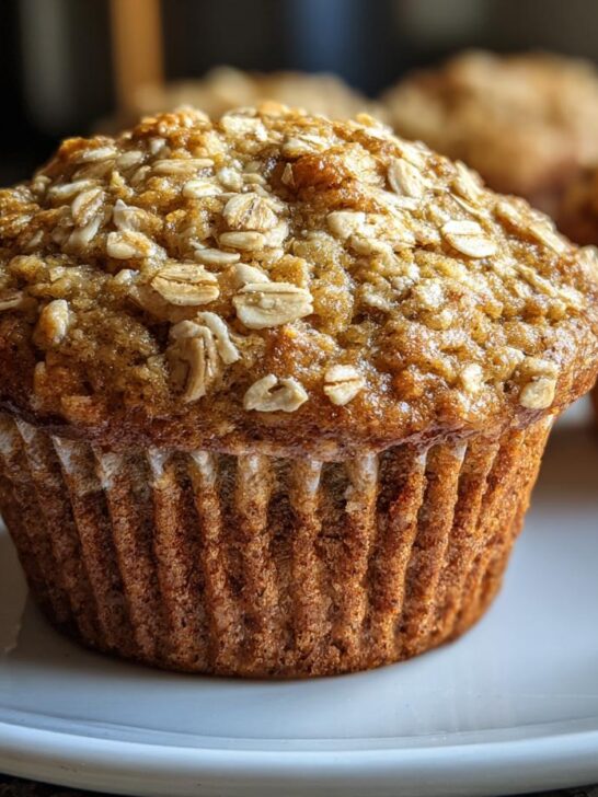 A close-up shot of a freshly baked Banana Oatmeal Muffin topped with visible rolled oats, sitting on a white plate.