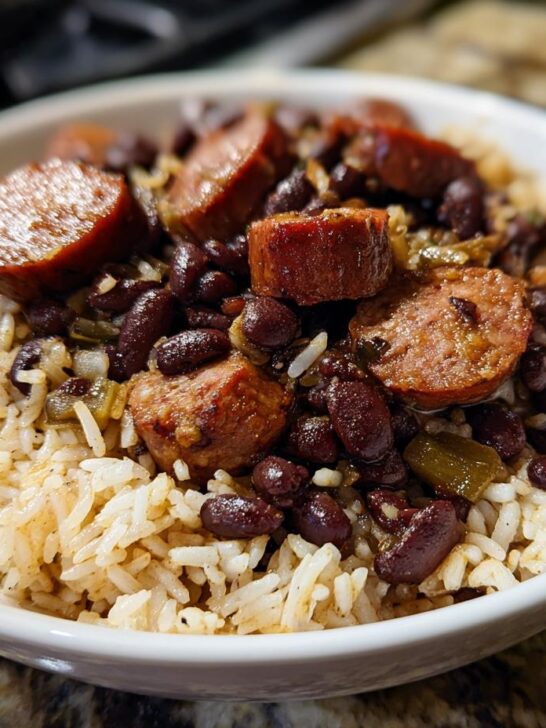 A white bowl filled with fluffy rice topped generously with dark red black beans and thick slices of browned sausage.