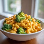 A close-up of a white bowl filled with creamy, cheesy Broccoli Mac And Cheese and bright green florets.