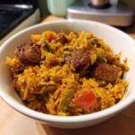 A close-up of a white bowl filled with flavorful Cajun Sausage And Rice Skillet, featuring browned sausage slices and colorful peppers.