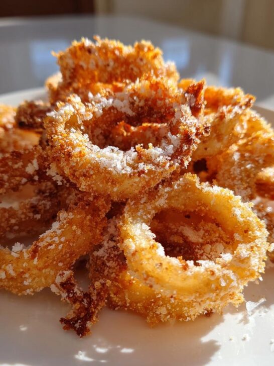 A close-up shot of crispy, golden Cheesy Onion Ring Chips piled high and dusted with white cheese or salt on a white plate.