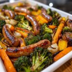 Close-up of browned chicken sausage and roasted vegetables like broccoli, carrots, and onions on a sheet pan.